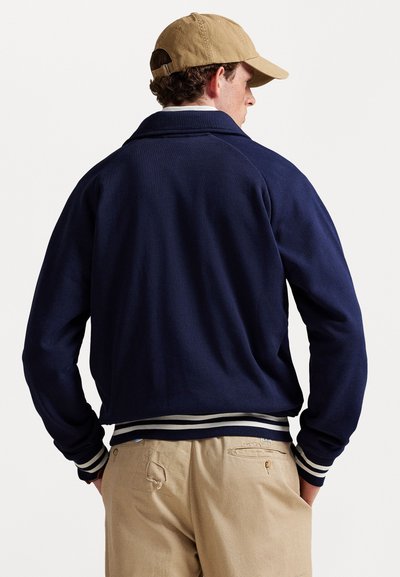 Uomo vestito con una giacca blu navy con polsini e cintura a strisce bianche, pantaloni beige con un strappo e un cappellino beige, di spalle.
