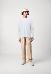 Light blue long-sleeve shirt paired with beige wide-leg trousers. The individual wears a beige cap and neutral sneakers. Simple, clean design.