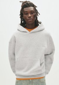 Lichtgrijze hoodie met een voorzak, een capuchon met trekkoorden en ribbelmanchetten. Gedragen over een oranje shirt, gecombineerd met lichtblauwe jeans.