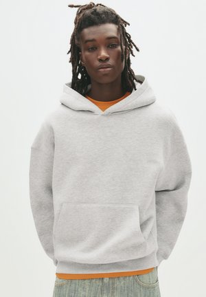 Lichtgrijze hoodie met een voorzak, een capuchon met trekkoorden en ribbelmanchetten. Gedragen over een oranje shirt, gecombineerd met lichtblauwe jeans.