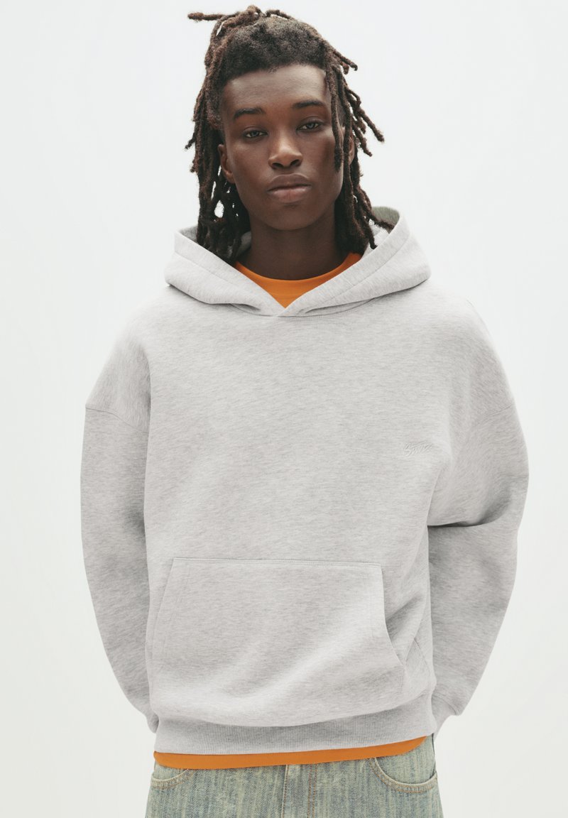 Lichtgrijze hoodie met een voorzak, een capuchon met trekkoorden en ribbelmanchetten. Gedragen over een oranje shirt, gecombineerd met lichtblauwe jeans.