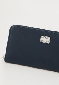 Portafoglio blu navy con superficie texture, chiusura a zip e una placca in argento con il logo "TOMMY JEANS" sul davanti. Design compatto e rettangolare.