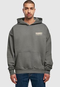 Grå hettegenser med kengurulomme. Har trykket teksten "HAWAI E KOMO MAI" i lys farge på brystet. Mykt stoff og avslappet passform.
