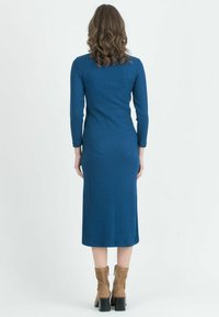 Donna con capelli ricci castani che indossa un vestito midi blu a coste con maniche lunghe e stivaletti con tacco color cammello, in piedi di spalle.