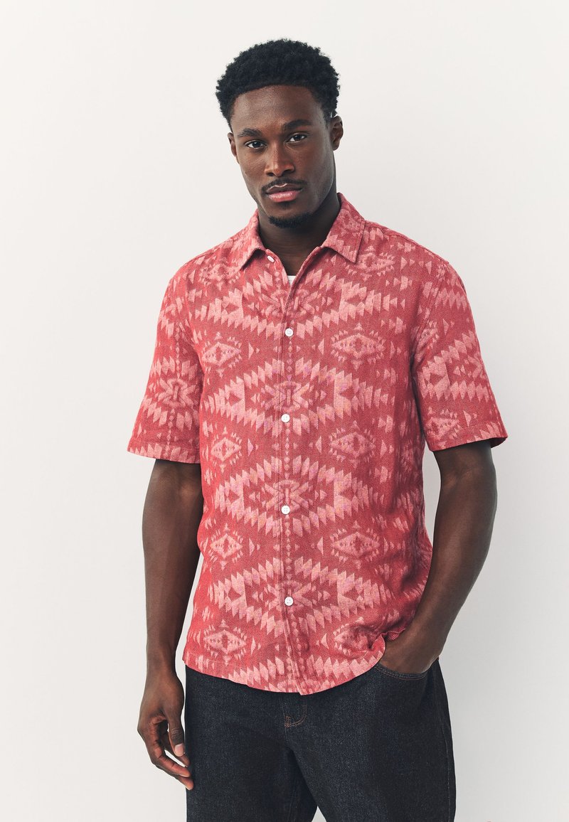 Camicetta rossa a maniche corte con motivo geometrico e dettagli bianchi. Realizzata in tessuto strutturato, presenta una chiusura frontale con bottoni e un colletto.