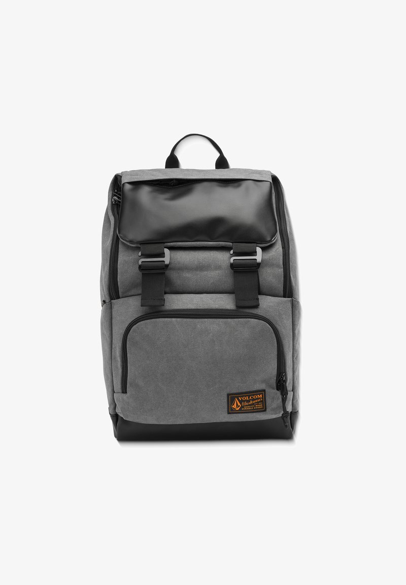 Sac à dos gris avec un rabat supérieur noir, bretelles réglables, poche zippée à l'avant et une étiquette logo orange. Fabriqué en tissu durable, design épuré.