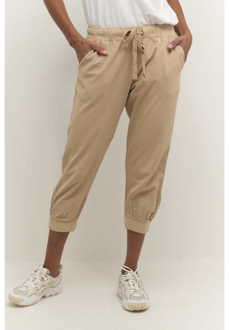 Persoană purtând pantaloni jogger bej croppați, cu manșete elastice și adidași albi, mâinile în buzunare, pe un fundal simplu.
