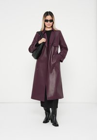 Purple faux leather coat with double-breasted button closure, side pockets, and a smooth texture. Worn over a black outfit and paired with black boots.