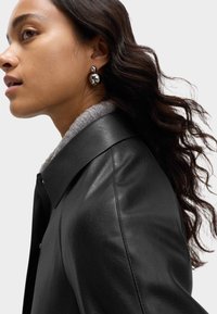 Černá kožená bunda s šedým límcem, se hladkým povrchem. Modelka má na sobě kulaté stříbrné náušnice a volné, vlnité tmavé vlasy.