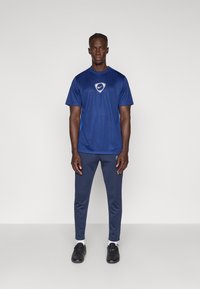 Ναυτικό μπλε αθλητικό t-shirt με κοντά μανίκια και κεντρικό λογότυπο· σε συνδυασμό με εφαρμοστά παντελόνια και μαύρα αθλητικά παπούτσια.