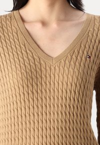 Pull col V marron clair avec un motif de tricot à cable texturé. Présente un petit logo de marque sur la poitrine gauche.