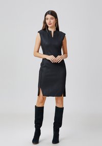 Robe noire sans manches avec un col montant, des fentes latérales et une texture lisse, associée à des bottes vert foncé montantes jusqu'aux genoux.