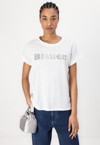 Camiseta blanca de manga corta con puños enrollados, que presenta un logo "Blauer." adornado con strass. Combinada con jeans de mezclilla azul y una bolsa gris.