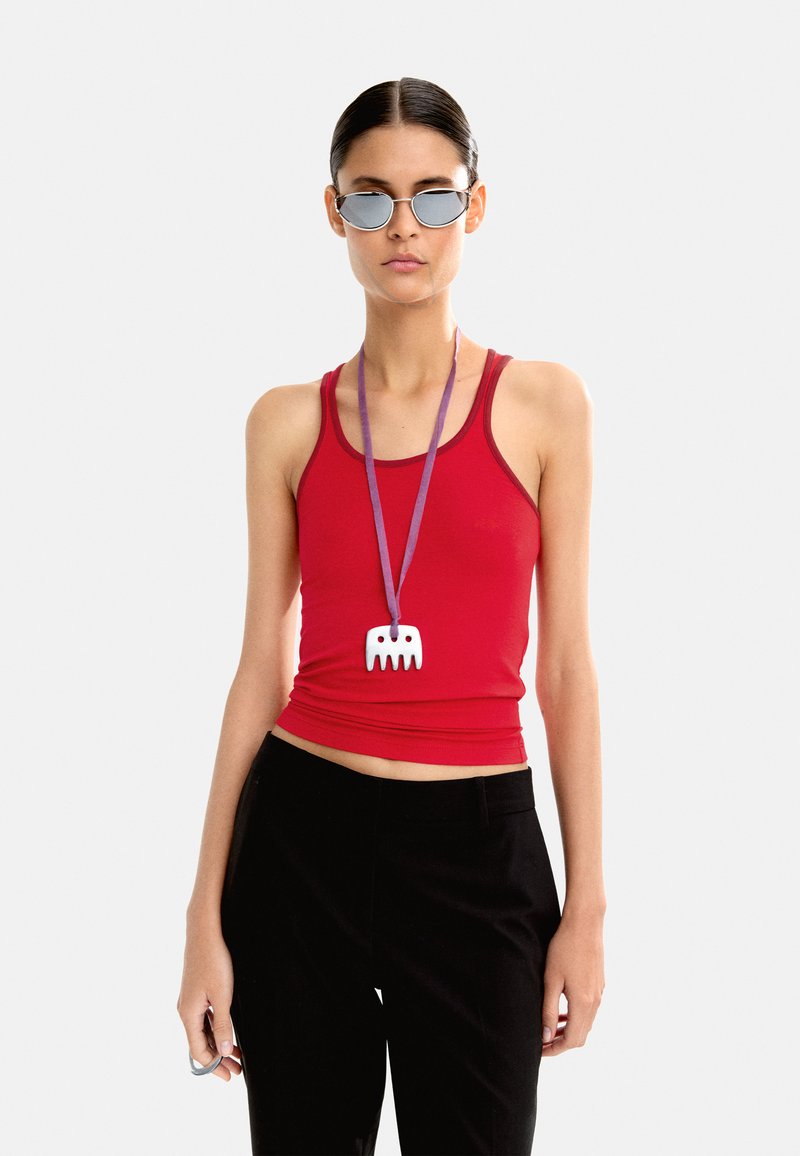 Czerwona dopasowana koszulka na ramiączkach, czarne spodnie z wysokim stanem, fioletowa smycz z białym wisiorkiem w kształcie grzebienia oraz okulary przeciwsłoneczne w srebrnym odcieniu. Gładkie białe tło.