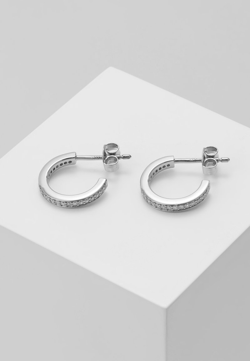 Paire de petites boucles d’oreilles créoles en argent avec des pierres transparentes incrustées, présentées sur une surface blanche inclinée.