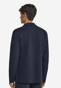 Homme portant une veste en tricot à manches longues bleu marine, vue de dos, avec un col rabattu et une finition droite.