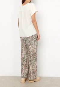 Blouse crème à manches courtes avec une couture au dos, portée avec un pantalon large à motifs paisley dans des tons verts, roses et gris.