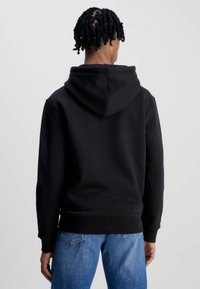 Felpa nera con cappuccio ampio, polsini e orlo a costine. Tessuto liscio, indossata con jeans azzurri chiari, vista da dietro.