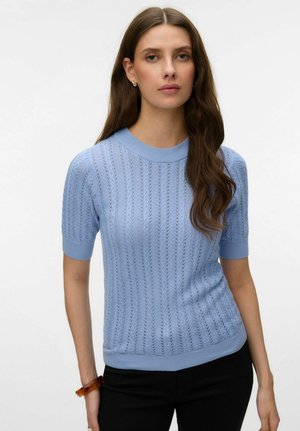 Femme aux longs cheveux bruns portant un pull tricoté à manches courtes bleu clair et un pantalon noir, debout devant un fond blanc uni.