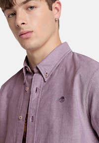 Camisa de botones de color morado claro con cuello, que presenta un logo en el pecho, acabada con costuras y botones en contraste.
