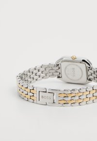 Montre en métal argenté et doré, avec un boîtier rectangulaire, un bracelet à maillons et un fermoir portant le logo gravé "BOSS". Finition lisse et accents polis.
