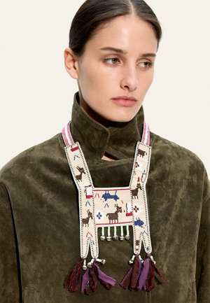Kobieta w oliwkowo-zielonej kurtce z zamszu i naszyjniku z koralikami z motywami zwierzęcymi, dzwoneczkami i fioletowymi frędzlami, na jasnym beżowym tle.