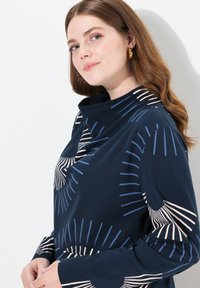 Marinblå tröja med hög krage, med ett solstråle-mönster i ljusblå och off-white. Mjuk tyg, avslappnad passform och långa ärmar.