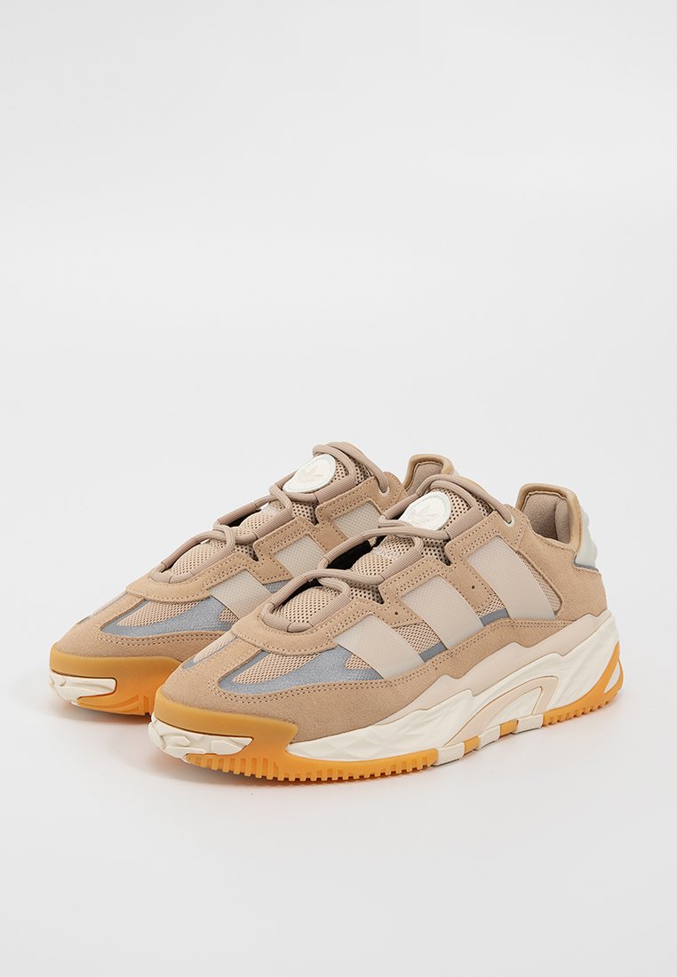 Beige Sportsneaker aus Mesh- und Wildledermaterial, mit drei seitlichen Streifen, strukturiertem Mittelsohle und orangefarbener Gummiaußensohle.