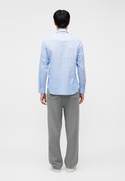 Άνδρας που στέκεται γυρισμένος με την πλάτη, φοράει μπλε ριγέ πουκάμισο, γκρι παντελόνι με τσέπες στην πλάτη και άσπρα αθλητικά παπούτσια σε απλό φόντο.