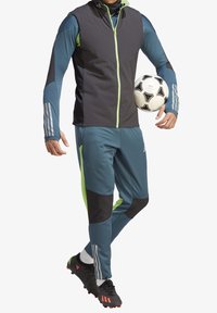 Conjunto deportivo que incluye una chaqueta de cremallera gris y verde azulado con acentos verdes, combinada con pantalones verde azulado y botas de fútbol negras, sosteniendo un balón de fútbol.