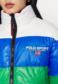 Femme portant une doudoune colorblock avec des sections blanches, bleues et vertes, brodée du logo "Polo Sport Ralph Lauren" et du drapeau américain.