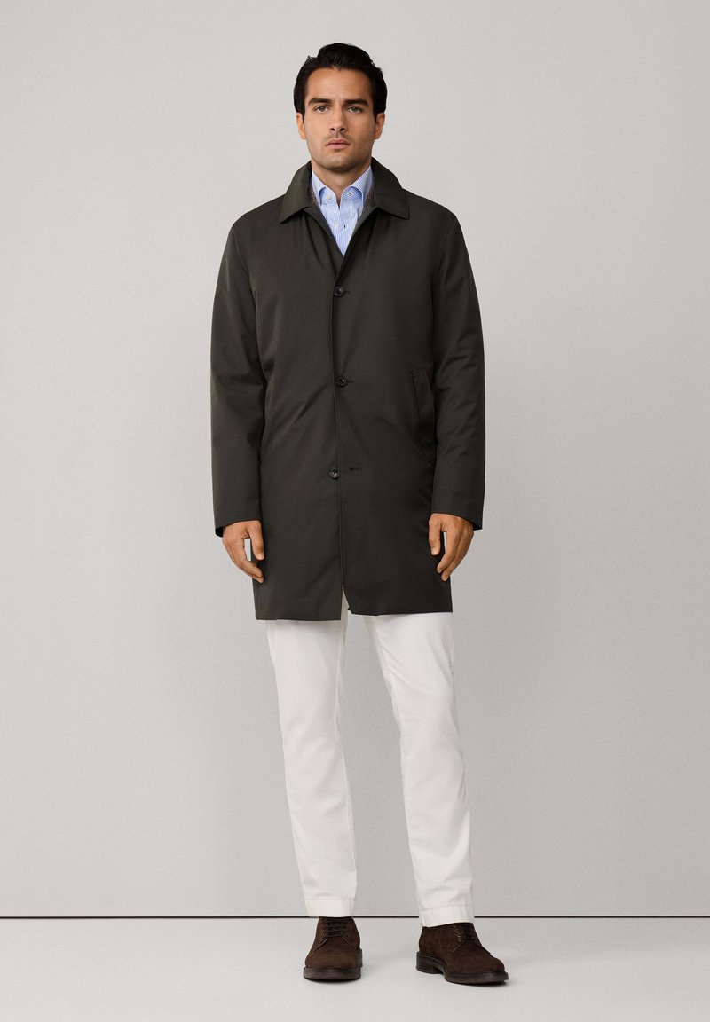 Tummanvihreä takki, jossa on kaulukset, etupainikkeet ja kaksi sivutaskua. Vaasattu sinisen nappipuseron päälle, yhdessä valkoisten housujen ja ruskeiden kenkien kanssa.