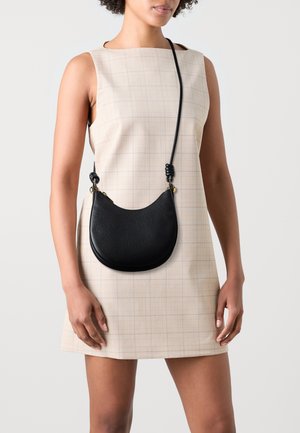 Sac bandoulière en cuir noir à la forme arrondie, surface texturée et détail de sangle nouée, porté sur une robe légère à carreaux.