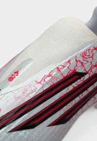 Ποδοσφαιρικό παπούτσι με γκρι επάνω μέρος που διαθέτει ανάγλυφο λουλουδένιο μοτίβο σε κόκκινο χρώμα. Μαύρες λεπτομέρειες περιλαμβάνουν τρεις ρίγες και κομψό σχεδιασμό.