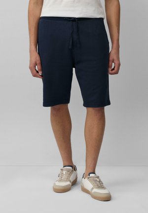 Shorts de survêtement bleu marine avec taille élastique et cordon de serrage, présentant des poches latérales et une coupe décontractée, assortis à des baskets beiges.
