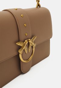 Brown leather handbag with a flap closure, gold-tone hardware, and a circular clasp featuring two birds. Accents include gold studs.