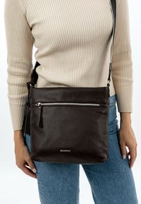 Brown leather crossbody bag with textured finish, front zip pocket, and brand logo. Adjustable strap and minimalist design.