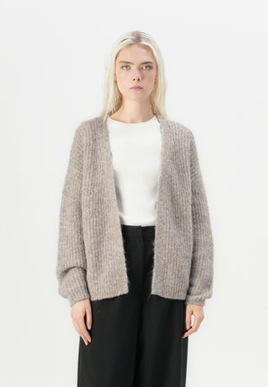 Beige strikket cardigan med ribbet tekstur, åpen front og lange ermer, kombinert med en hvit tettsittende skjorte og svarte bukser.