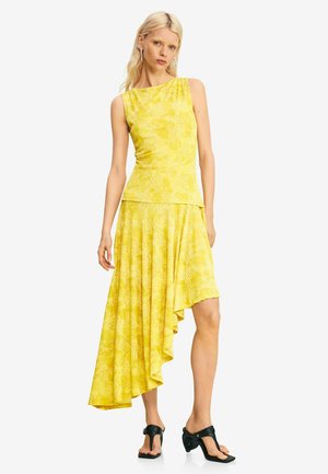 Blondínka v bezrukávovom žltom kvetinovom topu a asymetrickej žltej sukni s čiernymi sandálmi na opätku stojí na bielom pozadí.