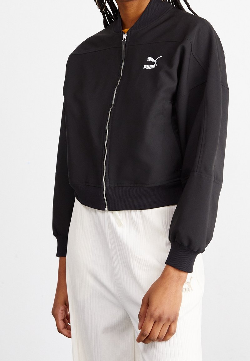Chaqueta bomber negra con cierre de cremallera, puños y dobladillo acanalados, con un logo de Puma blanco en el pecho izquierdo. Textura de tela suave.