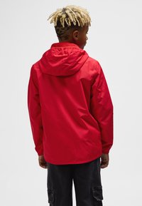 Red waterproof jacket with a hood, featuring elastic cuffs and a loose fit. Smooth texture with a plain design, worn over black pants.