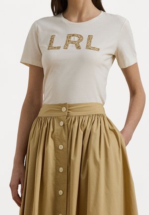 Vrouw draagt een beige T-shirt met korte mouwen met gestructureerde "LRL" letters en een hooggetailleerde lichte bruine rok met knopen aan de voorkant.