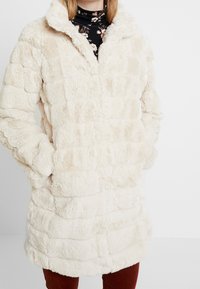 Konstpälsjacka i krämfärg med en strukturerad, flätad design. Har en hög krage, långa ärmar och sidofickor. Mjuk och plyschig material.