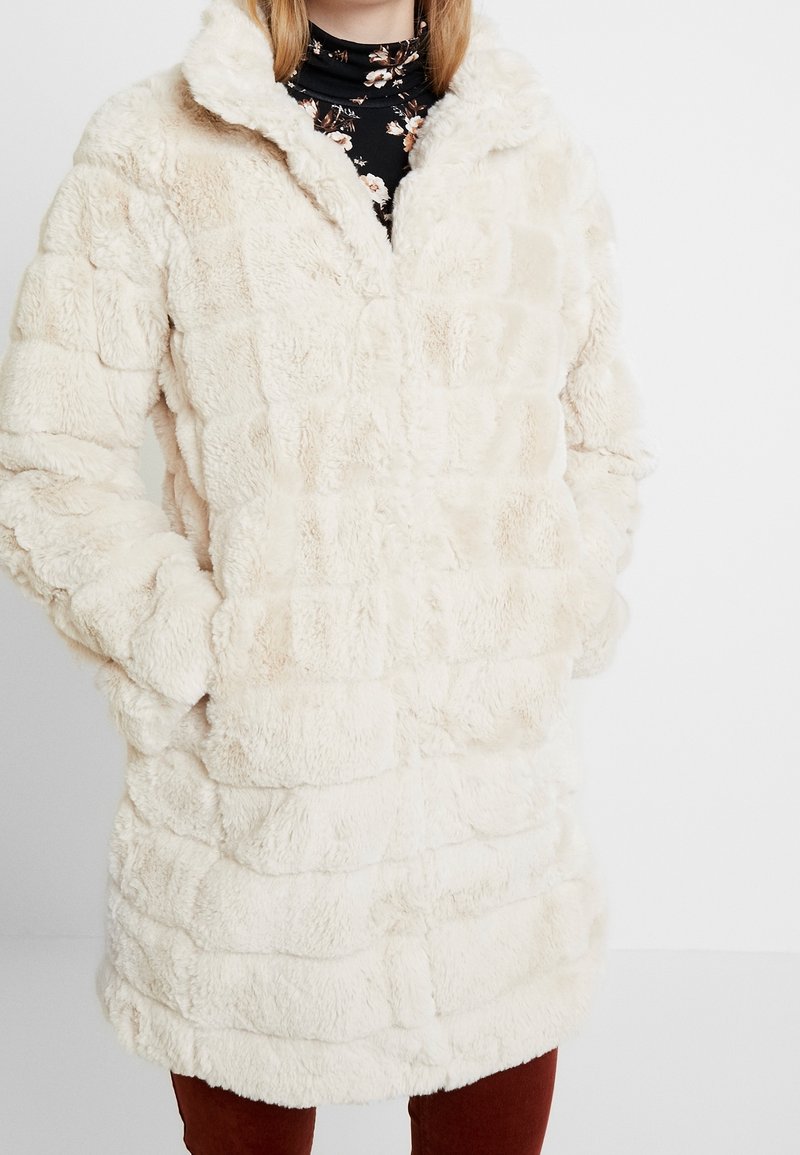 Konstpälsjacka i krämfärg med en strukturerad, flätad design. Har en hög krage, långa ärmar och sidofickor. Mjuk och plyschig material.