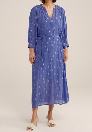 Femme portant une robe midi bleue à manches longues avec des accents verticaux argentés, une ceinture à la taille et des sandales blanches à talons avec des détails floraux.