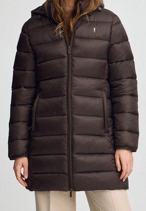 Vrouw draagt een donkerbruine doorgestikte lange donsjas met ritssluiting aan de voorkant, zijzakken en een klein geborduurd logo op de borst.