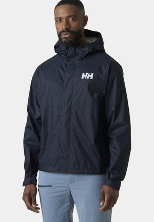 Hombre que lleva una chaqueta impermeable con capucha de color azul marino con el logo "HH" en el pecho y pantalones azul claro, de pie contra un fondo liso y claro.