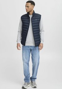 Mornarsko pufasta jopica, lahka, z zadrgo spredaj, nošena čez sivi pulover s kratkimi rokavi, v kombinaciji s svetlimi denim hlačami in sivimi supergami.