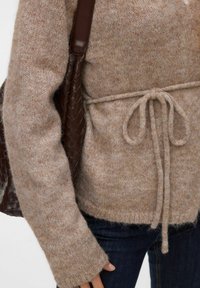 Smeđi pleteni džemper s dugim rukavima, vezanim strukom i mekanom teksturom. Uz to, smeđa ispletena torba na ramenu.