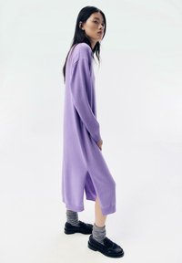 Langes, lila gestricktes Kleid mit lockerem Schnitt, seitlichen Schlitzen und gerippten Bündchen, kombiniert mit grauen Socken und schwarzen Schuhen.